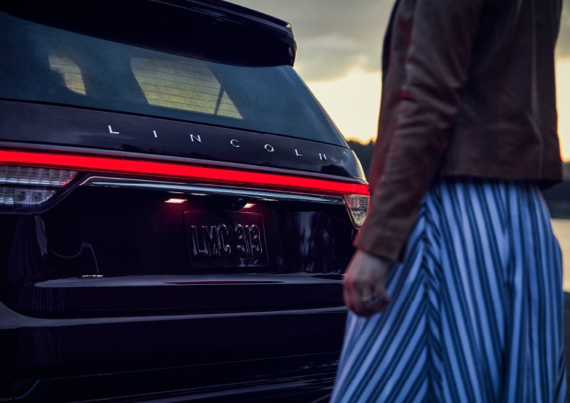 A person is shown near the rear of a 2024 Lincoln Aviator® SUV as the Lincoln Embrace illuminates the rear lights | Varsity Lincoln in Novi MI
