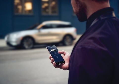 A person is shown interacting with a smartphone to connect to a Lincoln vehicle across the street. | Varsity Lincoln in Novi MI