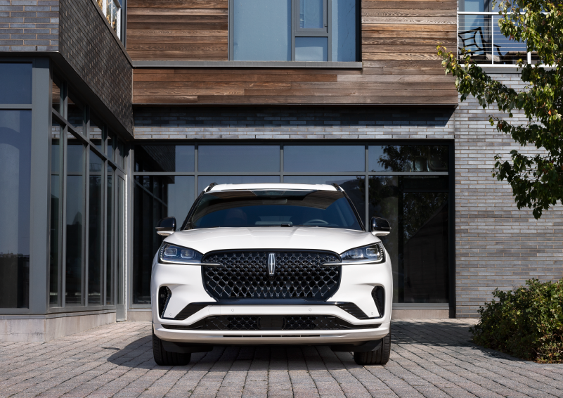 A white 2025 Lincoln Aviator® SUV is shown parked outside a modern building with gray brick and wood accents. | Varsity Lincoln in Novi MI
