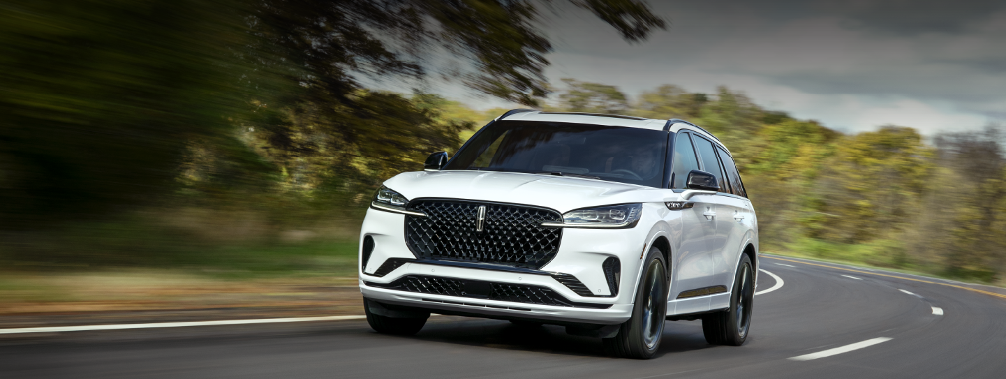 A white 2025 Lincoln Aviator® SUV is being driven down a road lined with trees. | Varsity Lincoln in Novi MI