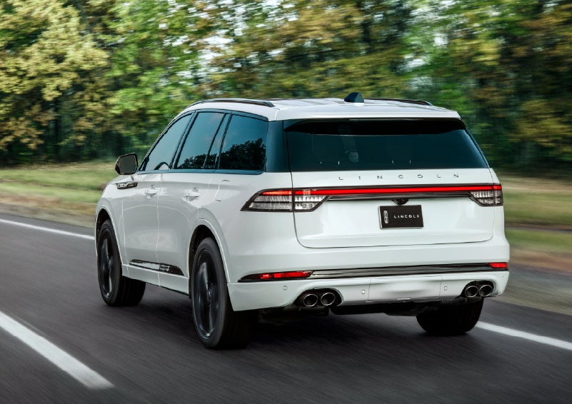 A white 2026 Lincoln Aviator® SUV is being driven away down a road lined with colorful trees. | Varsity Lincoln in Novi MI